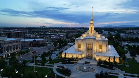 LDS-Mormon-Temple-in-Ogden-Utah-drone-flight-flying-at-dusk-on-beautiful-summer-night-static-slightly-angled-shot-as-water-fountain-and-American-flag-blowing-in-breeze-and-cars-driving-by-in-the-city