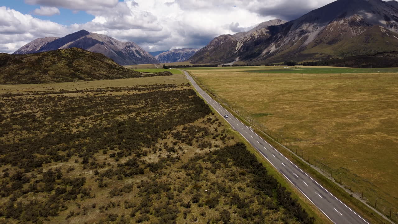 Wide aerial drone shot of a car driving along State Highway 73 in New Zealand’s Island Alps near Arthur’s Pass. Flat road, high scenic view with Purple Hill, dramatic peaks, and alpine landscape