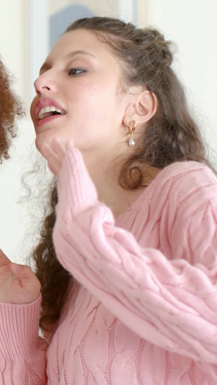 Vertical video: Leaning left as friend talks, woman in pink sweater blowing kiss to greet at home