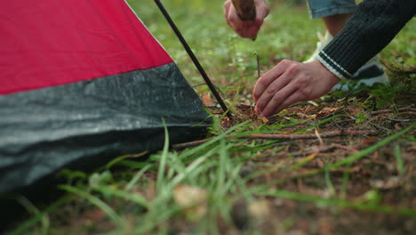 close up partial view of person crouching in forest using short wood stick to hammer metal tent peg into ground beside red tent surrounded by grass dry pine needles and branches