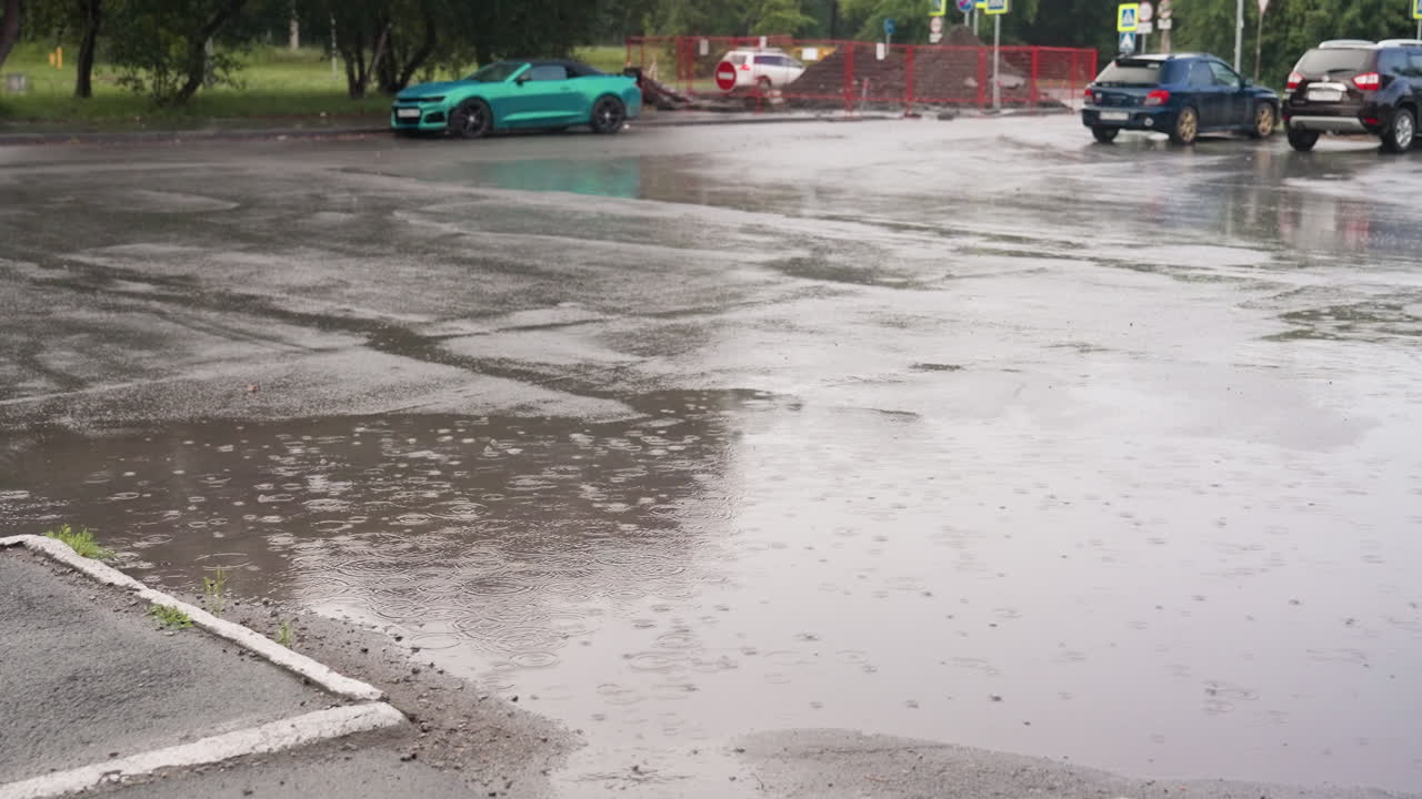 Wet ground filled with rainwater forming puddles as cars remain parked nearby, rippling reflections on surface capture rainy atmosphere and urban detail with vehicles surrounding flooded asphalt area