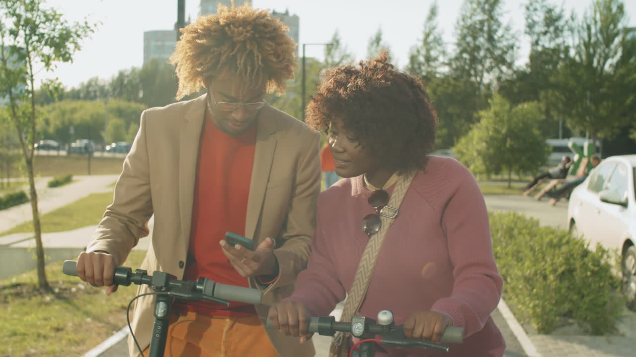 Amigos afro-americanos alegres parados no parque com patinetes elétricos e usando o celular
