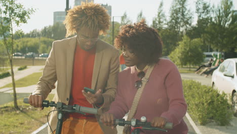Cheerful-African-American-Friends-Standing-in-Park-with-E-Scooters-and-Using-Phone