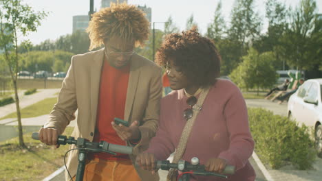 cheerful african american friends standing in park with e-scooters and using phone