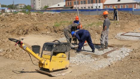 Form-workers-work-on-the-layout-of-foundations-on-a-construction-site,-a-worker-walks-past-with-a-wheel-barrow