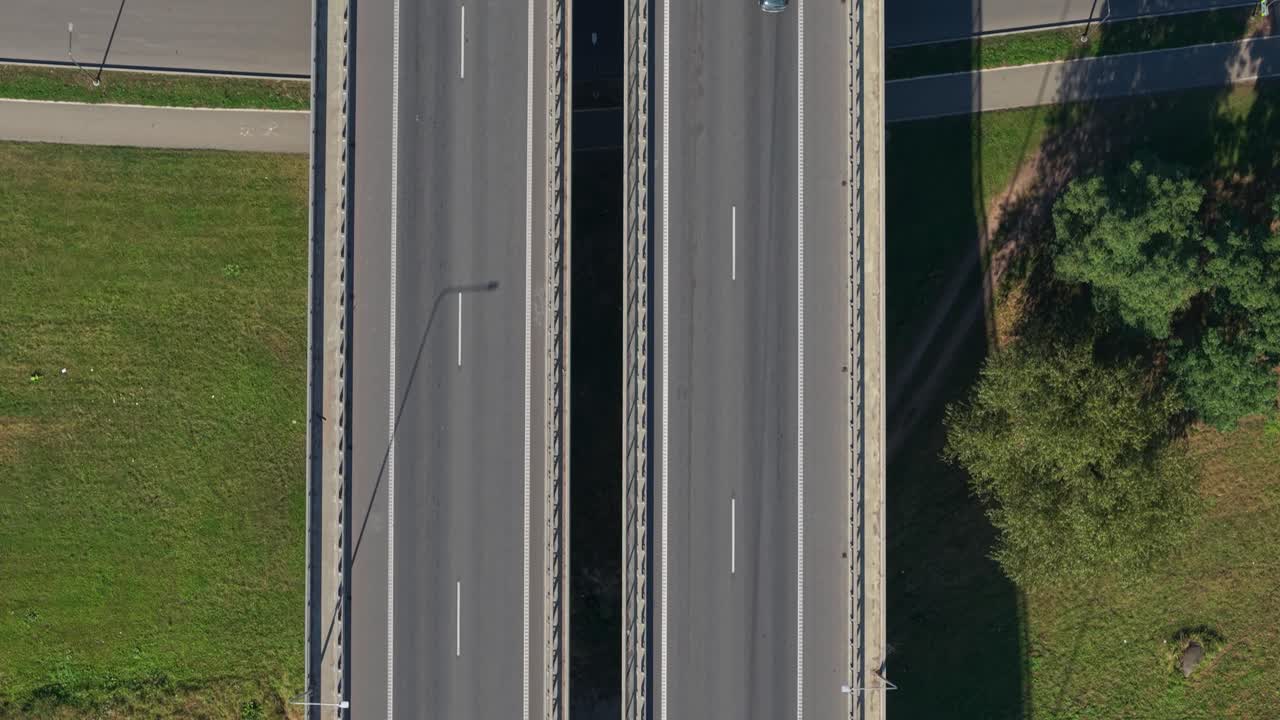 Top-down aerial view of a highway bridge with moving cars and a freight truck crossing over a river in Kaunas, Lithuania. Clear weather and sharp vehicle shadows enhance the scene