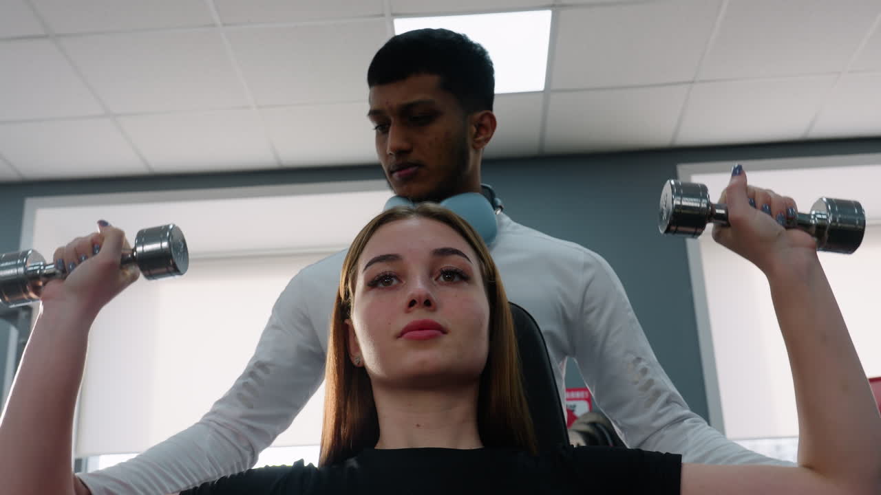 trainer in white top with headphones guides young woman through overhead dumbbell press in bright gym setting with focused expression and strength form instruction from experienced coach