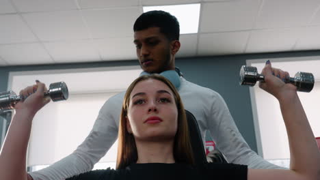 trainer in white top with headphones guides young woman through overhead dumbbell press in bright gym setting with focused expression and strength form instruction from experienced coach