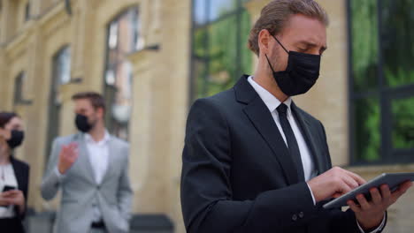 Masked-businessman-using-tablet-outdoors.-Suit-man-maintaining-social-distance