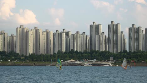 Many-People-Windsurfing-on-Han-River-Daytime,-Jamsil-apartments-on-background,-cloudy-day,-stunning-colorful-sunset-in-Seoul