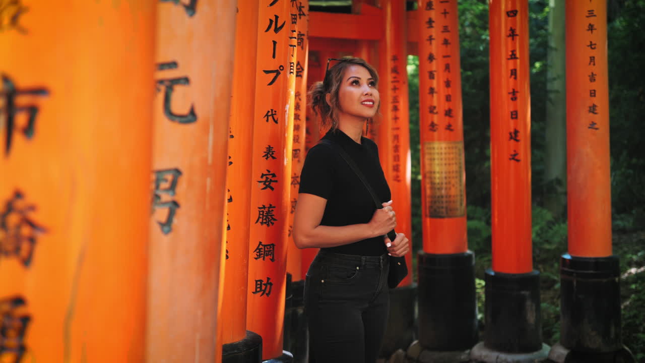 женщина с кудрявыми светло-коричневыми волосами, одетая в черный топ и джинсы, наслаждается своим визитом в храм фушими инари в ярко-оранжевых воротах тори с японскими надписями создают живописный фон