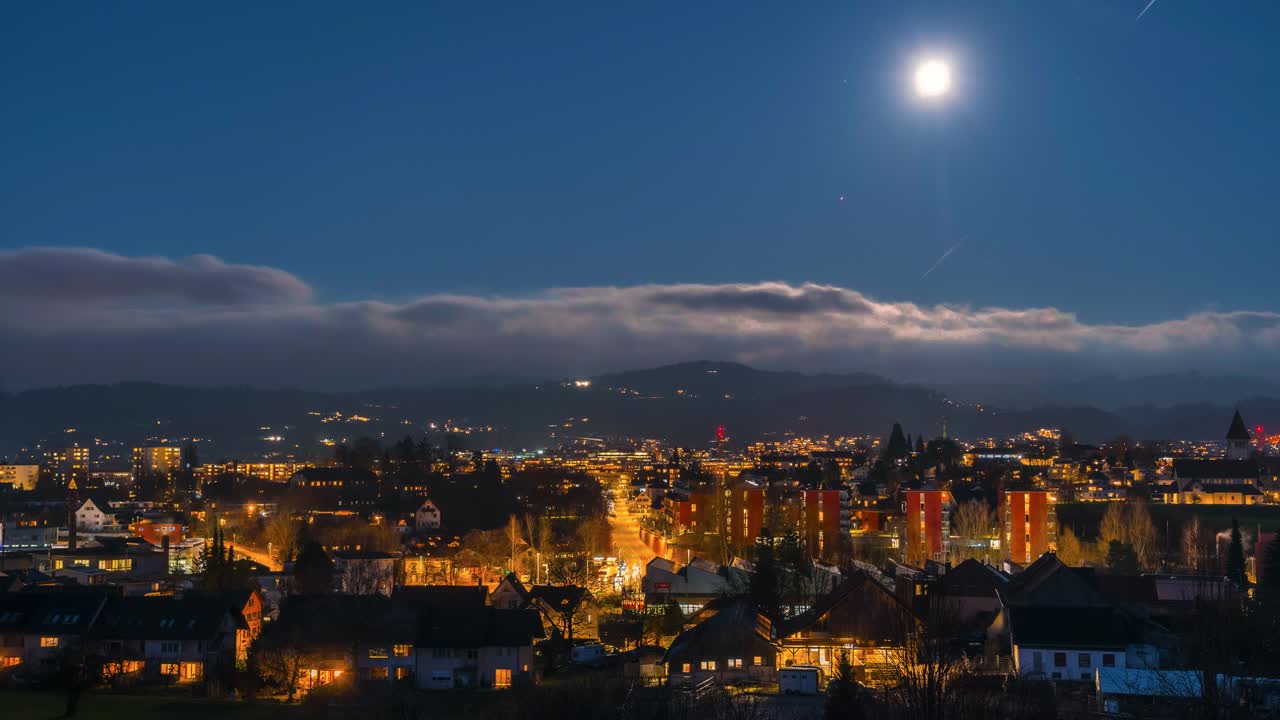 Time lapse full moon over a city with lights and car tracks. The clouds move on the horizon and the lights illuminate the city. There are small mountains in the background.