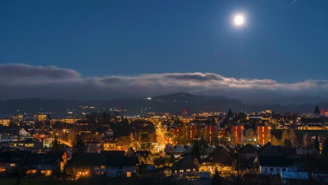 time lapse full moon over a city with lights and car tracks. the clouds move on the horizon and the lights illuminate the city. there are small mountains in the background.