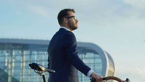 vista lateral de un apuesto hombre de negocios con traje y gafas, caminando y sosteniendo su bicicleta fuera de un edificio moderno de la ciudad