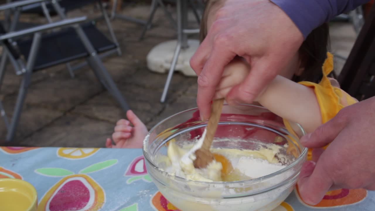 작은 아이와 어머니의 중간  ⁇ , 케이크 혼합을  ⁇ 는 데 도움이, 야외 테이블에 앉아, 낮 동안