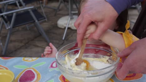 Mid-shot-of-small-child-and-mother,-helping-to-stir-cake-mix,-sat-outside-at-patio-table,-during-the-daytime