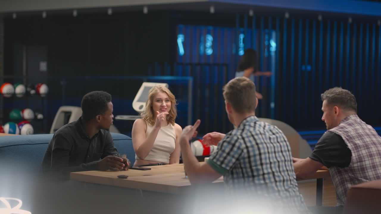 Um grupo multiétnico de amigos em um clube de boliche conversa à mesa, sorrindo, celebrando e trocando high-fives com um amigo que fez um strike. Homem e mulher negros