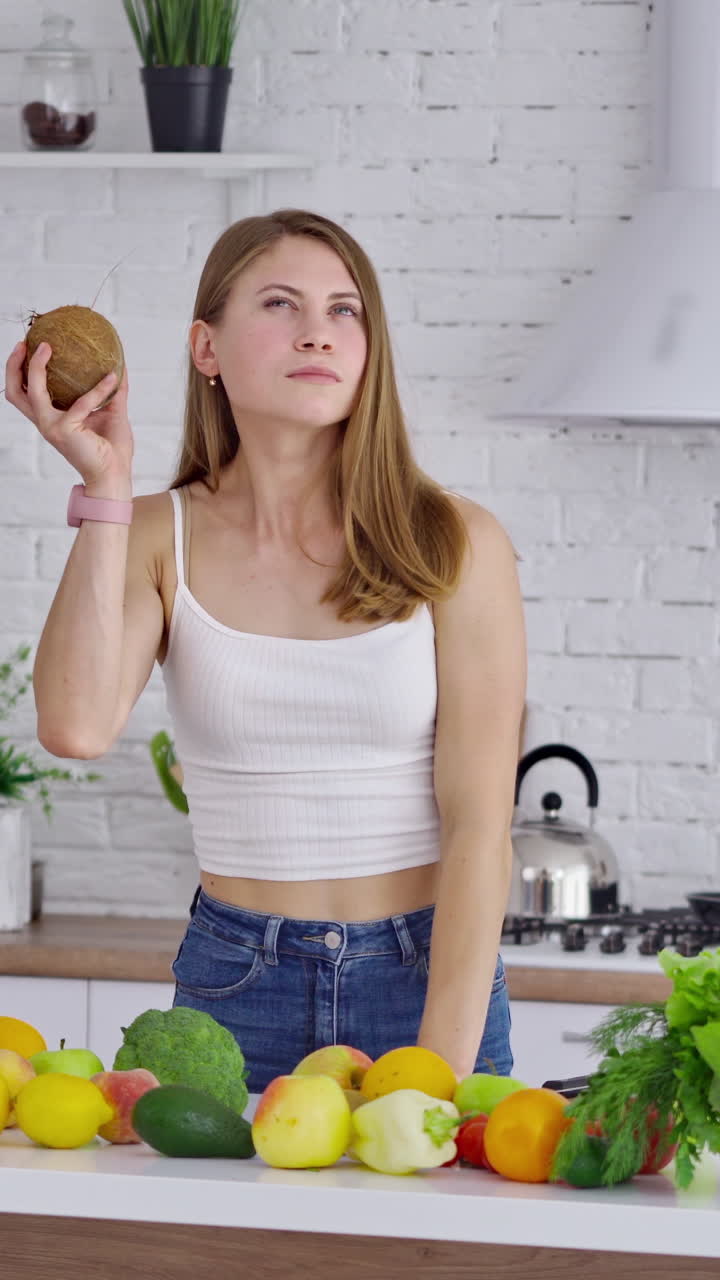 Beautiful woman with coconut after shopping. Young lady holding a fresh fruit and smiling on the kitchen background. Fresh fruits and vegetables on the table. Healthy food concept. Vertical video
