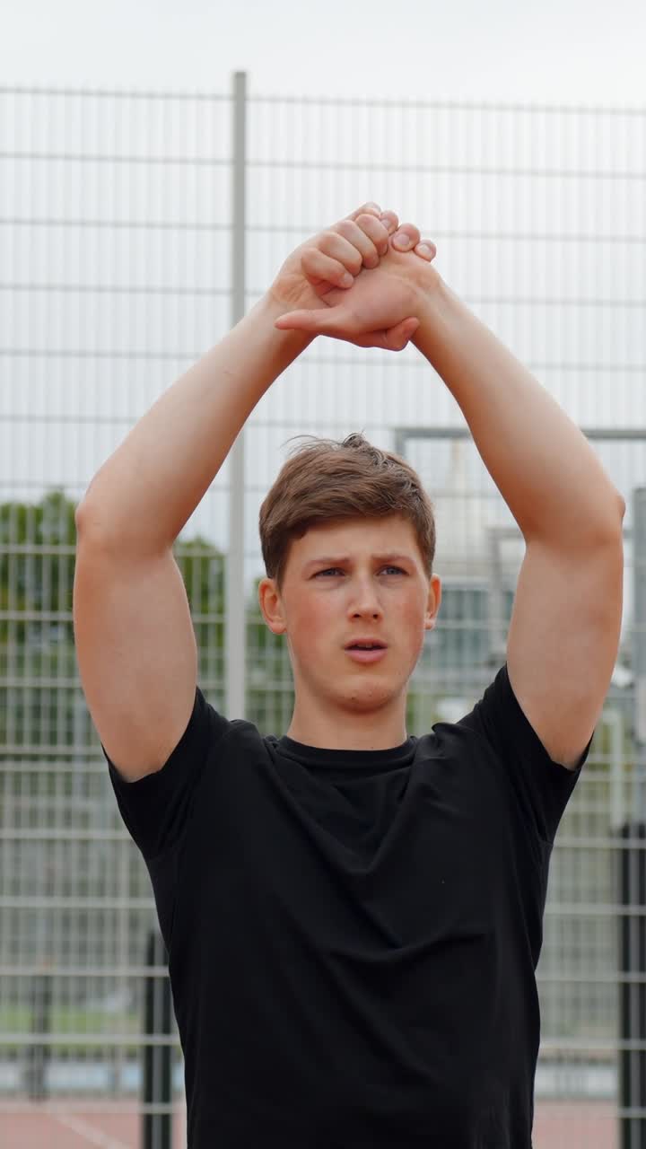 Young man standing outdoors, performing an arm stretch by raising his clasped hands above his head, focusing on his warm up exercise with a determined expression, vertical static, slow motion shot