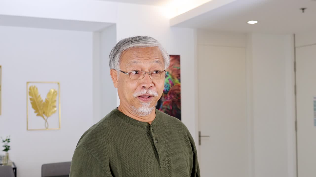 Senior Asian man smiles and points, cheerful expression, well-lit modern home interior, static camera