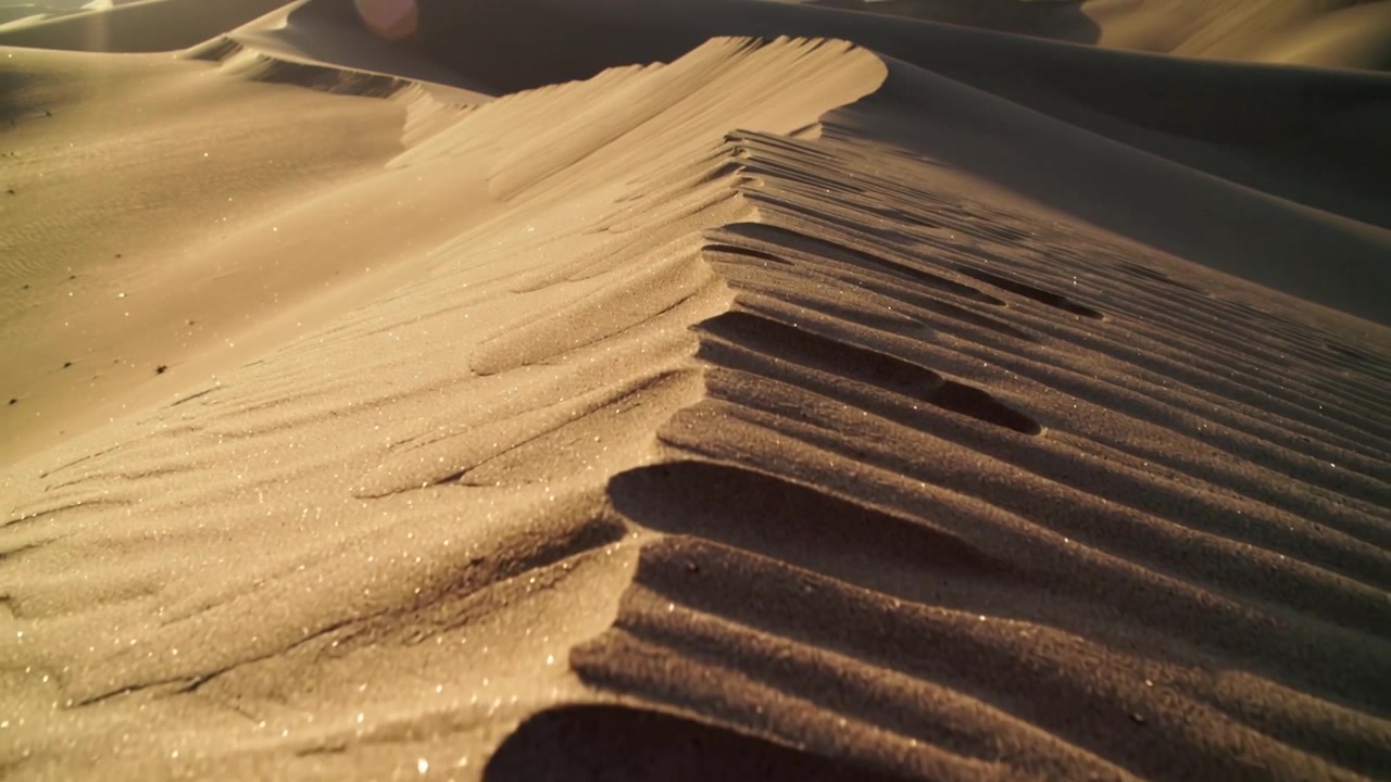 Telecamera che si sposta filmando la cresta di una duna di sabbia nel deserto, catturando la texture, i granelli scintillanti e le increspature.