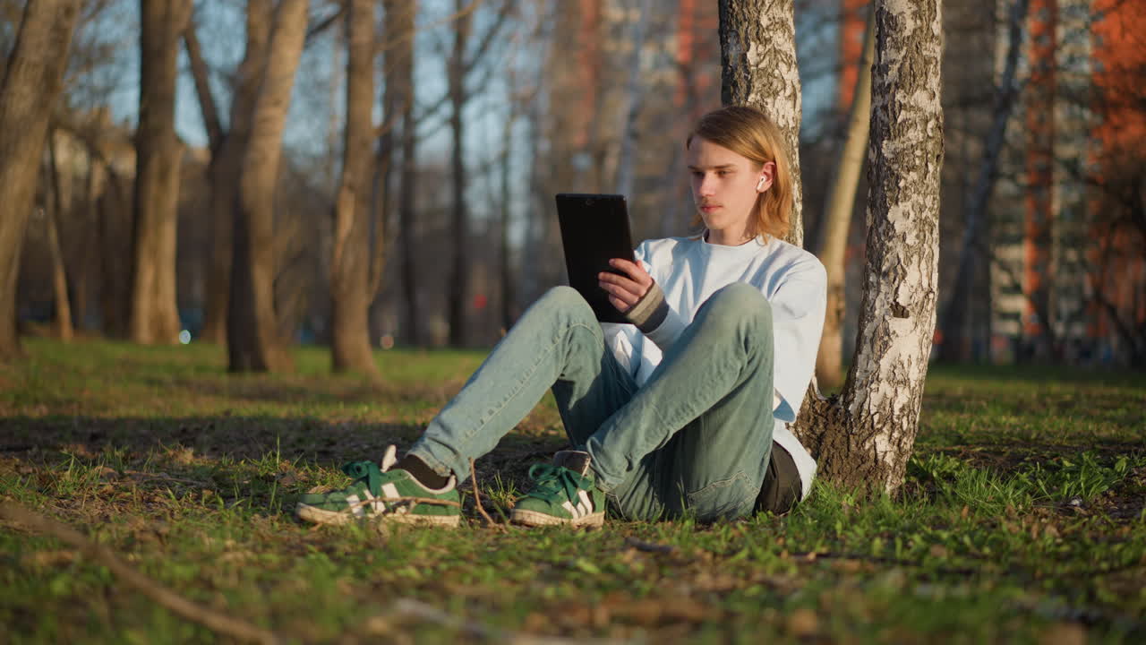 adolescente blanco leyendo una tablet apoyado en un abedul en un parque, con las piernas dobladas sobre el césped y una mochila cerca; tranquila sesión de estudio a la luz cálida de la tarde, estudiante concentrado disfrutando del aire fresco