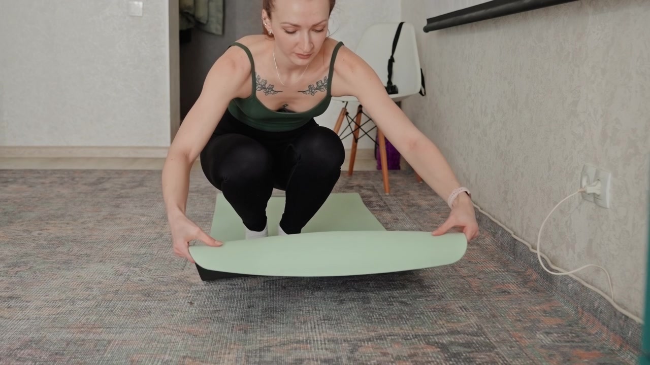 Female arranges mat calmly indoors. Woman unrolls mat in living area for fitness practice with focus. Female calmly prepares her yoga mat at home to engage in exercise session peacefully