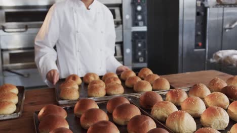 Portrait-of-happy-asian-female-baker-in-bakery-kitchen-with-fresh-rolls-on-counter-in-slow-motion