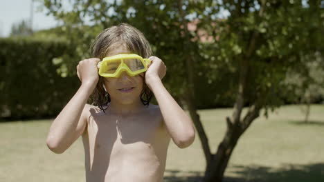 vista frontal de un niño feliz con el torso desnudo y gafas para deportes acuáticos mirando la cámara en el jardín de casa