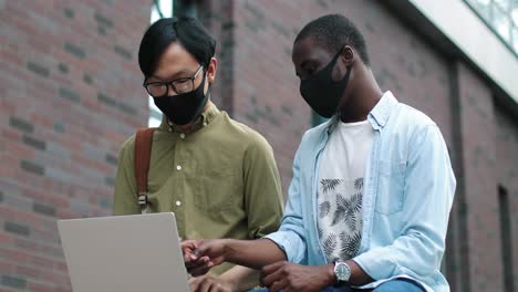 waist up portrait view of asian and african american male students wearing protective masks sitting in the street and looking at the laptop while studying new the college