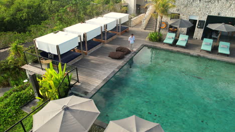 young man walks along swimming pool and sits on beanbag at tropical luxury hilltop villa in bali indonesia, wide aerial