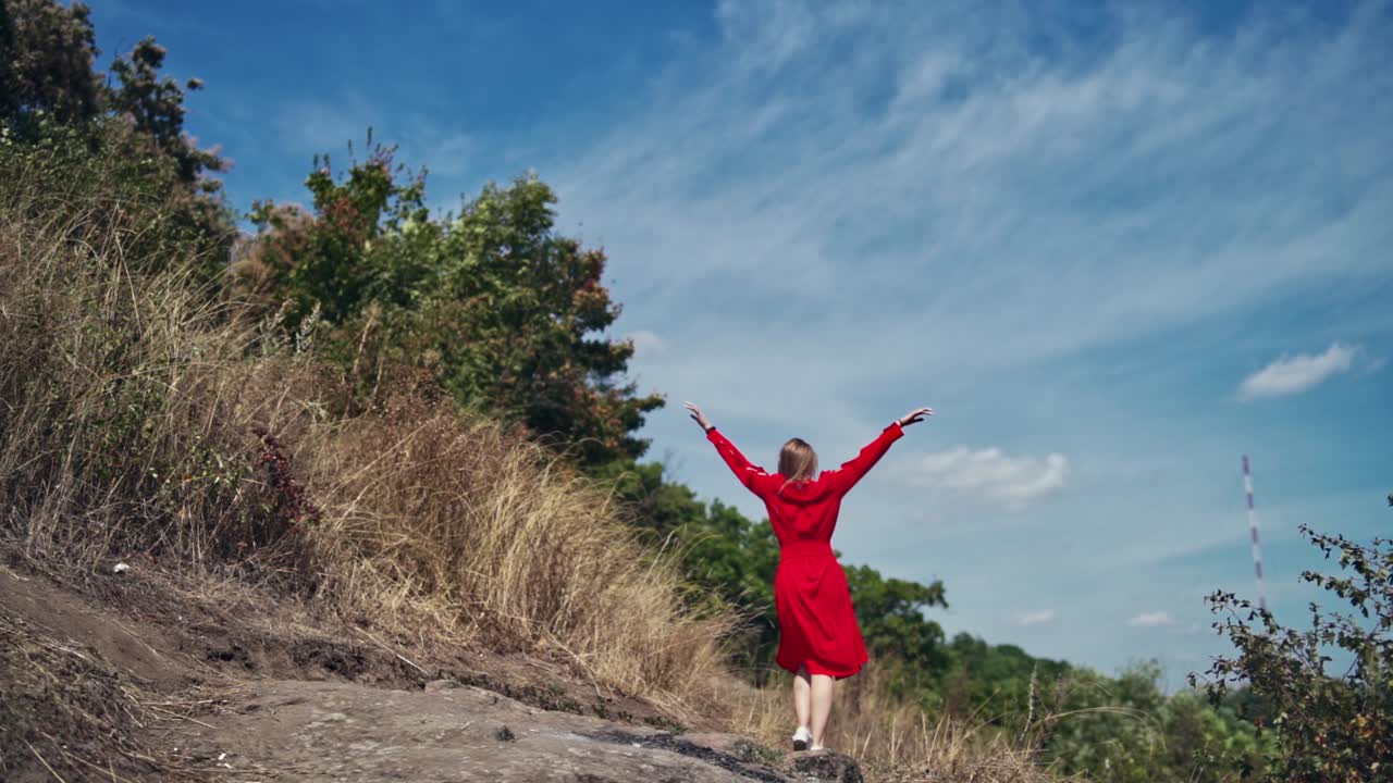 Backside view of young woman outdoors. Happy woman in beautiful red dress walking among nature. Lady enjoys summer landscape.