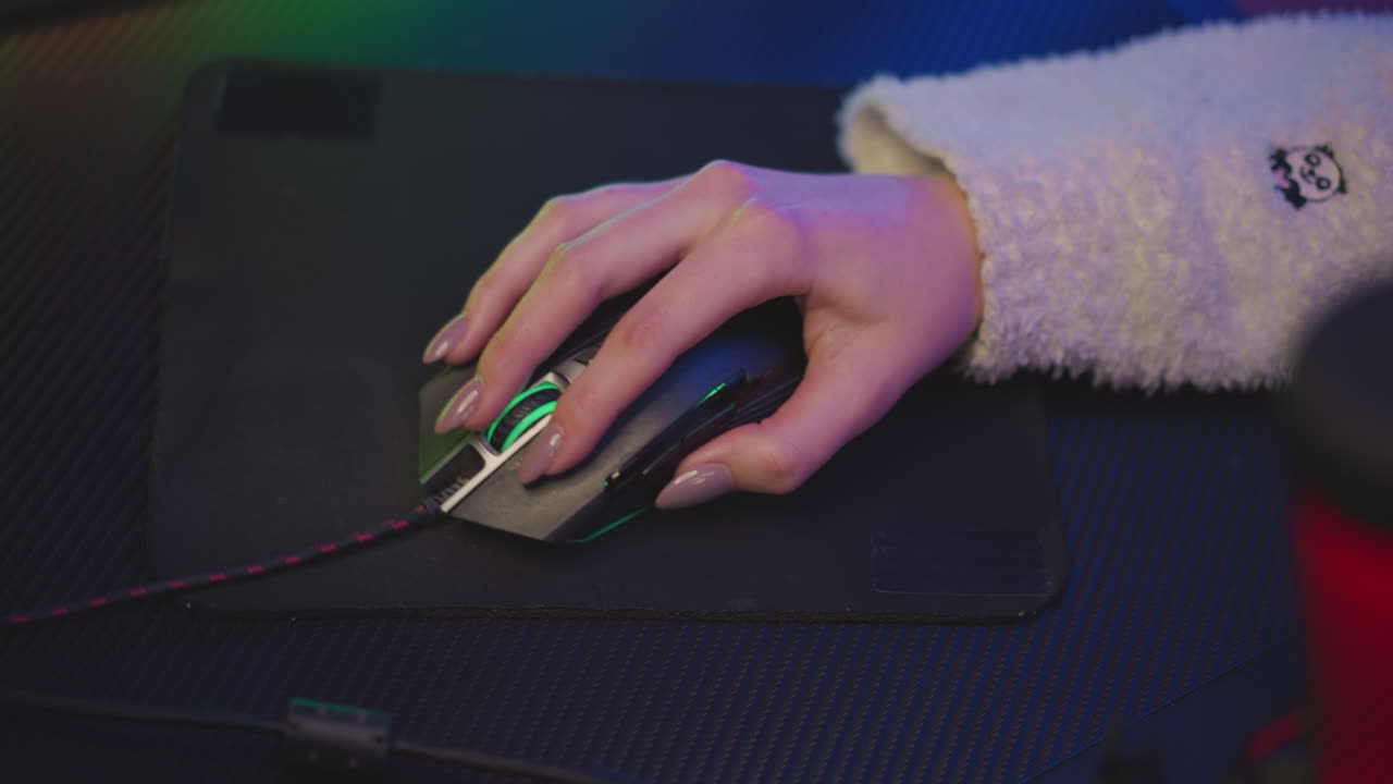 Woman with polished nail in fluffy teddy bear sweater using gaming mouse on textured black pad, lit by subtle pink and green ambient lighting from LED strip and microphone