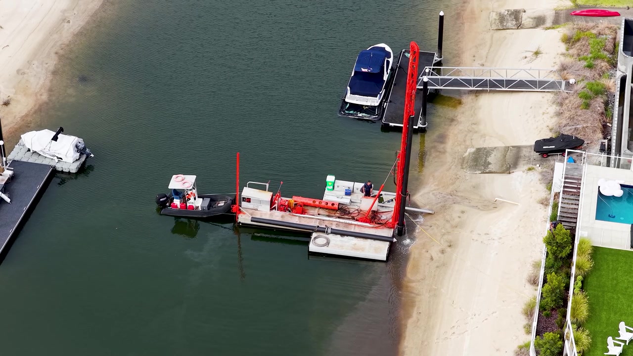 Vista aerea di attrezzature industriali per il dragaggio in funzione vicino a proprietà residenziali di lusso fronte mare in Australia.