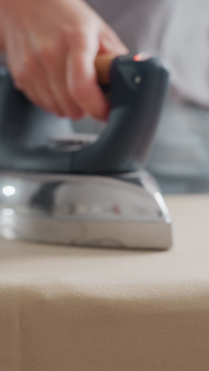 closeup white woman professional presser using steam station, confident hand guiding iron over beige fabric, commercial technique with docked appliance, laundry technician smoothing large textile
