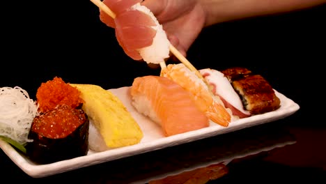 a hand skillfully picks assorted nigiri sushi with chopsticks from a neatly arranged platter, under dramatic studio lighting with a black background