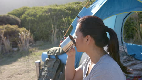 camping in mountains, woman drinking from metal cup near tent and backpack
