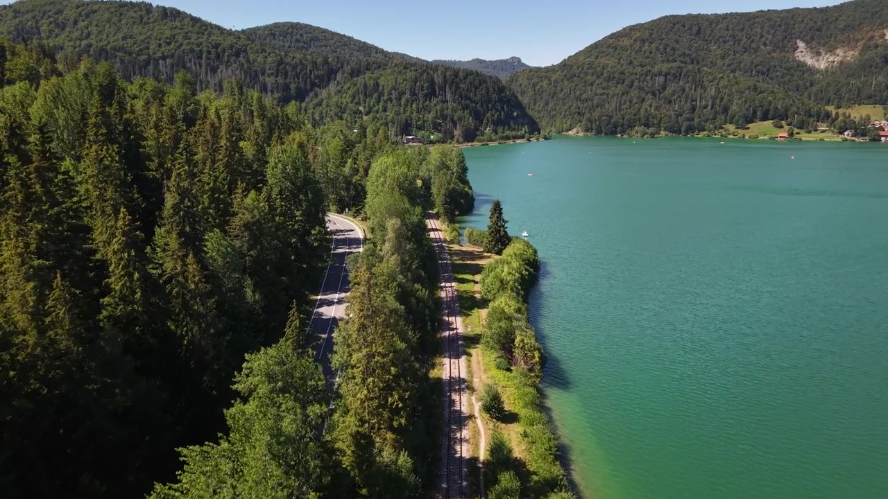 An aerial view captures a tranquil turquoise alpine lake, bordered by lush green forests, mountains, railway tracks, and a parallel road.
