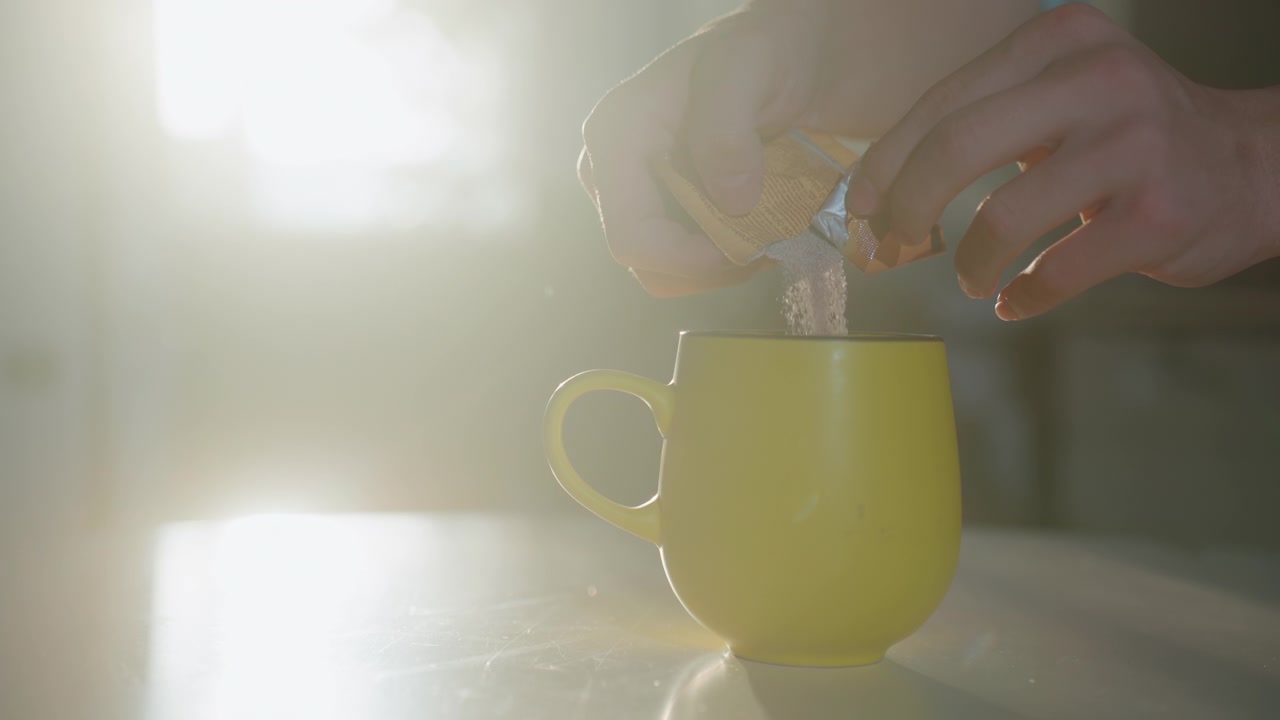 Mani illuminate dal sole che versano un preparato istantaneo in una tazza gialla, retroilluminazione delicata, primo piano delle dita che strappano la bustina, granuli che cadono a cascata nella tazza, vapore leggero, tavolo da cucina, rituale intimo della colazione, cinematografico
