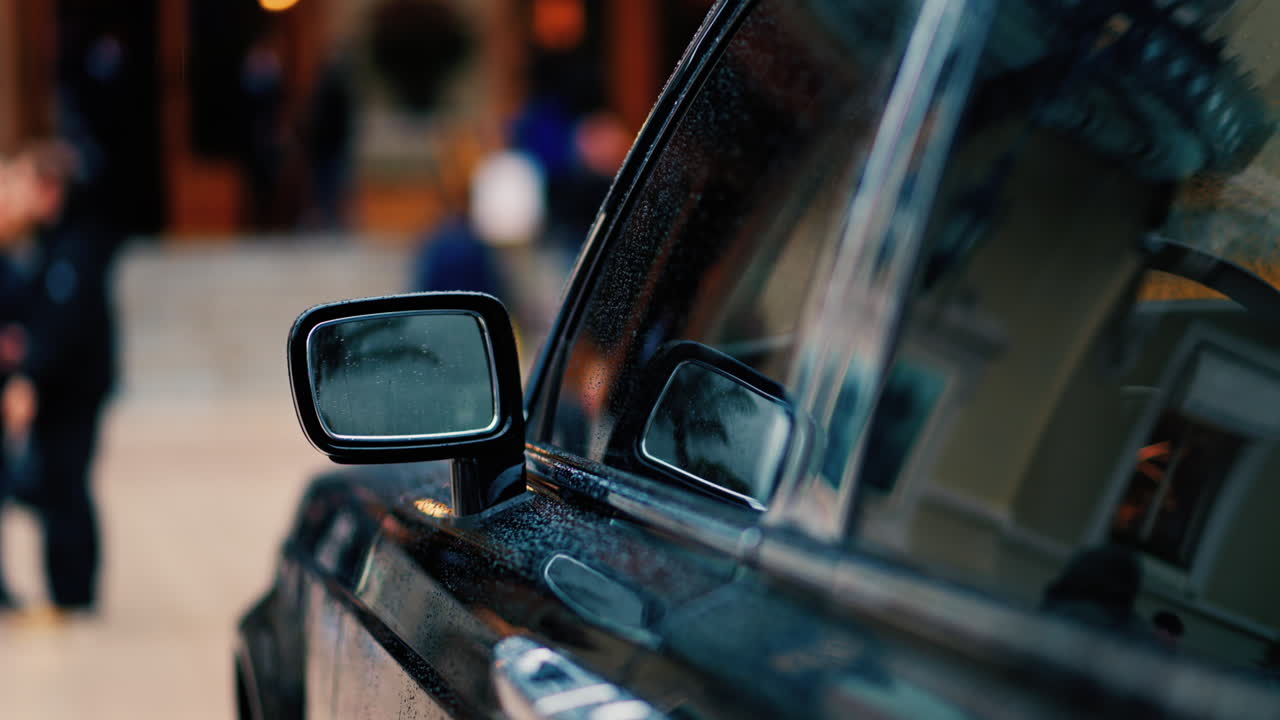 Close up of the mirror of a black car in the rain with people walking on a blurred background