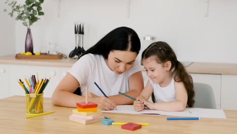 beautiful mom and her pretty daughter sitting at table in kitchen and drawing together