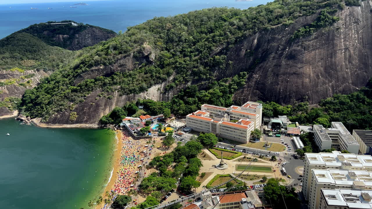 Uma vista panorâmica ampla para a direita do Morro da Urca, mostrando a movimentada Praia Vermelha, o histórico prédio do IME e a exuberante Mata Atlântica que circunda os picos de granito do Rio