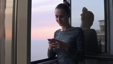 Joven-Mujer-Atractiva-Enviando-Un-Mensaje-De-Texto-Mientras-Está-De-Pie-Junto-A-La-Ventana-Abierta-Con-Una-Sonrisa-Durante-La-Puesta-De-Sol-Junto-Al-Mar