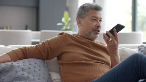 un hombre de dos razas está hablando en su teléfono inteligente sentado en el sofá en la sala de estar, en cámara lenta.