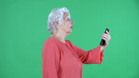 retrato de una mujer anciana hablando en una llamada de video usando su teléfono inteligente. abuela de cabello gris en blusa roja en la pantalla verde en el estudio. vista lateral. primer plano