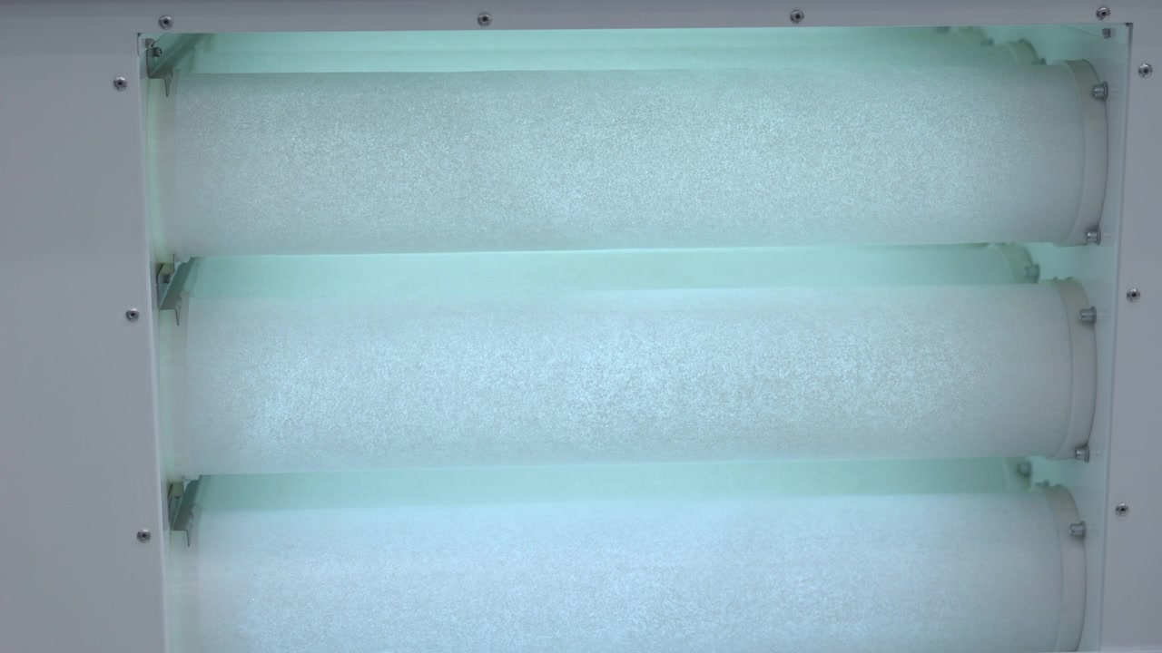 Vista ravvicinata di apparecchiature di filtrazione industriale con tre elementi filtranti cilindrici all'interno di un alloggiamento metallico chiaro, illuminato da una fredda luce ciano che evidenzia superfici texturizzate, bulloni, staffe e.