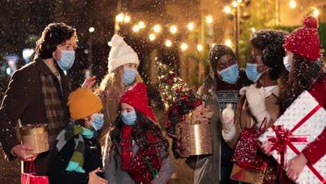 fröhliche multiethnische freunde und kinder unterhalten sich und halten geschenke auf der straße, während es zu weihnachten schneit