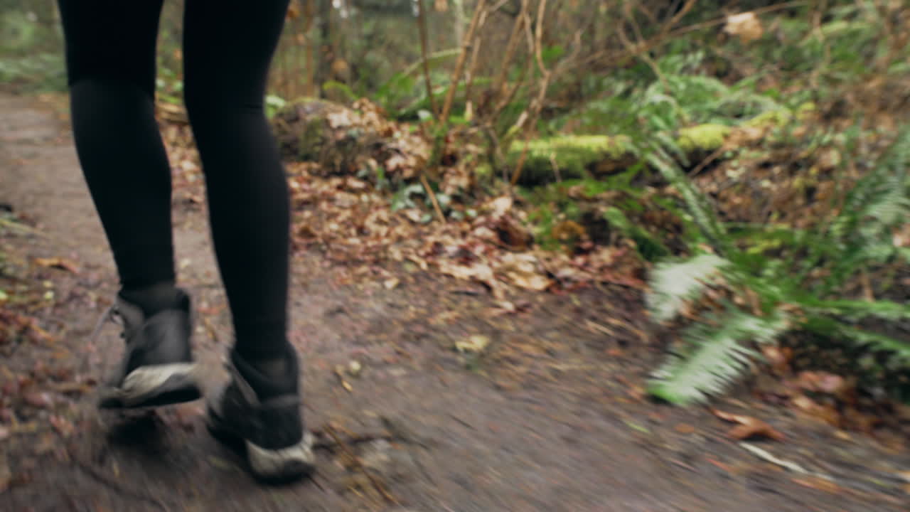 A young woman wearing a bright yellow jacket walks down a boardwalk in a green mossy forest, tracking shot of her boots