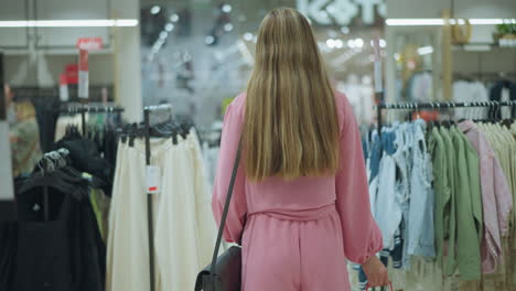 back view of a lady in pink with hand black bag carrying shopped items, walks through a clothes rack in a brightly lit shopping mall, other shoppers are blurred in the background, observing clothes