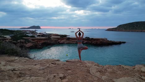 vista aérea mágica vuelo de arriba posición de chica de yoga árbol atardecer acantilado playa isla de ibiza españa drone ascendente